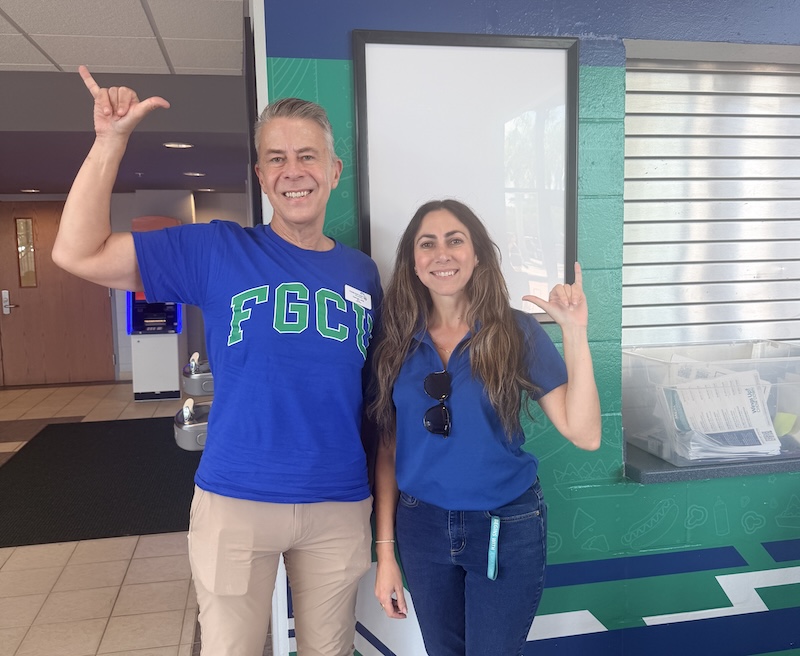 Two individuals stand inside Alico Arena and pose for a photo while making the Wings Up salute. One person wears a blue FGCU T-shirt, and the other wears a blue polo shirt. A blue-and-green wall and a counter are visible in the background.
