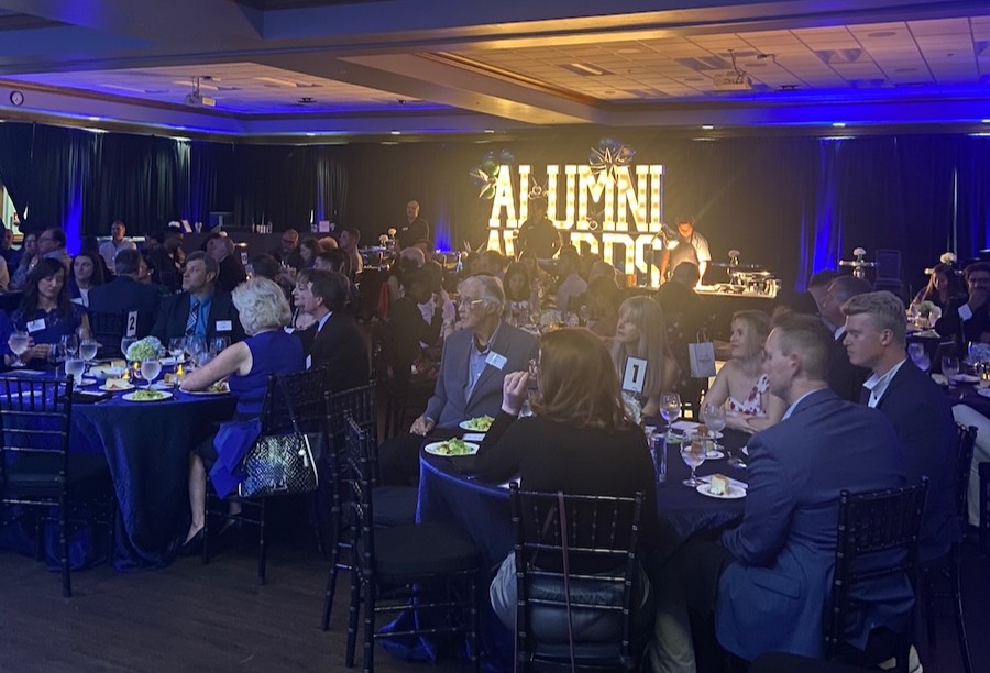 Dressed in business casual attire, guests at the Alumni Award celebration are sitting at round tables enjoying dinner. The venue features dim blue lighting throughout the room and an LED light display in the words “Alumni Awards” is displayed in the background along with a catering table.