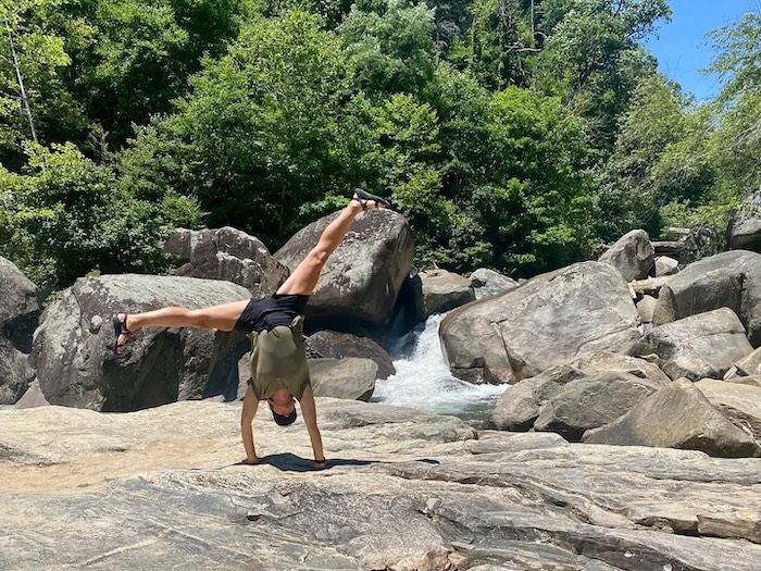 Amanda Whittamore is performing a handstand on a rocky surface near the Green River in North Carlina. Large boulders and lush green trees are in the background. 