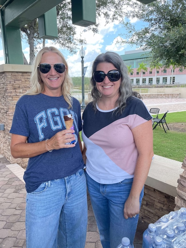 Two people stand side by side under a pavilion. One person wears a blue Ģ��TV T-shirt and holds a frozen dessert, while the other wears a color-blocked shirt. Both are wearing sunglasses. 