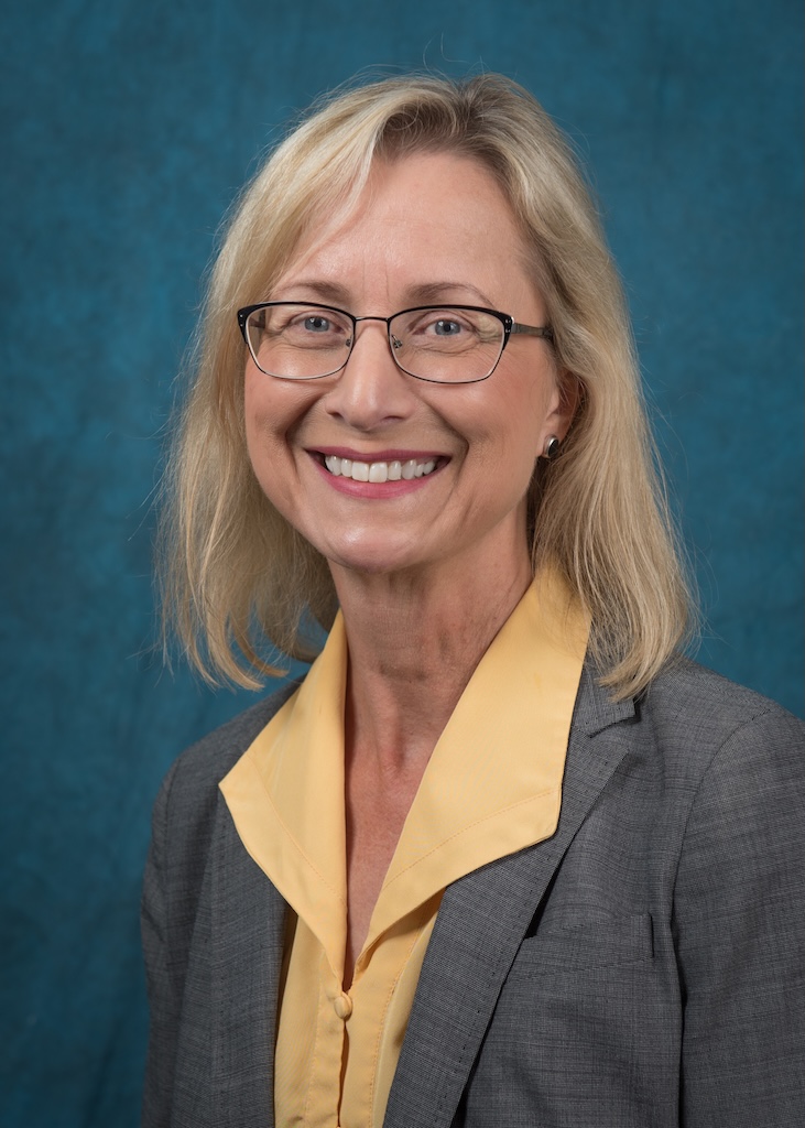 Headshot of Ann Rose. She has short blonde hair, and is wearing glasses and a yellow shirt underneath a grey jacket.