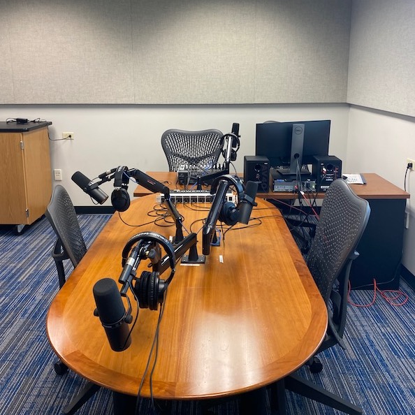 A recording studio featuring a wooden table with four mounted microphones and headphones. Two office chairs surround the table. In the background, a desk holds a computer monitor, audio equipment and cables. The room includes carpeted flooring and acoustic panels on the walls.  