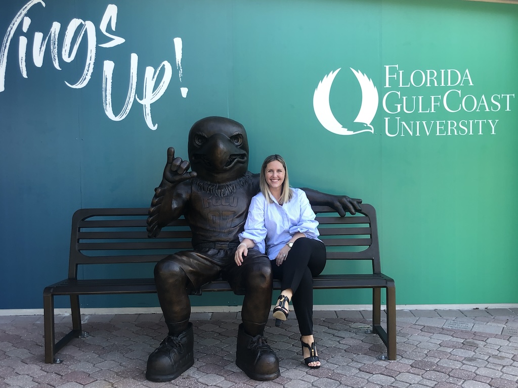 Wearing a blue and white button-down shirt, black pants, black open toe shoes and a watch on her left wrist, Kimberly Wallace, senior director of Alumni Relations, is sitting on a bronze bench with a bronze statue of FGCU’s mascot Azul. The bench is placed in front of a green and blue gradient wall and on top of octagon shaped outdoor tile.  