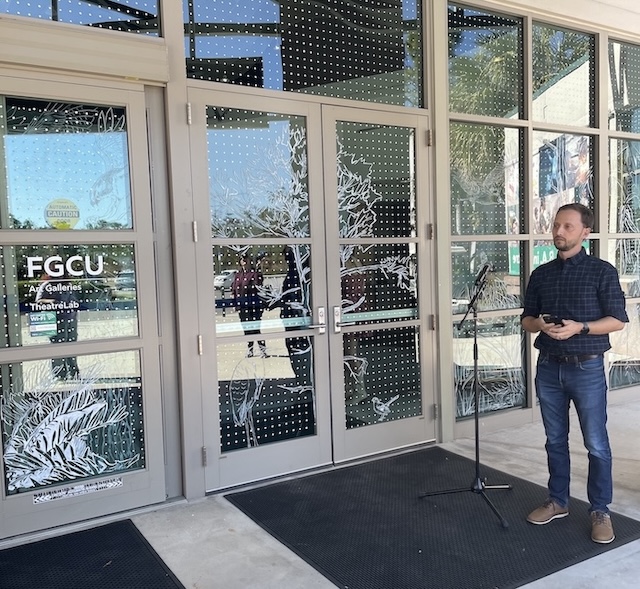 Professor Oscar Johnson stands outside the entrance of the FGCU Arts Complex next to the main entrance glass doors. They are decorated with a white-dot pattern and white nature‑inspired designs. A microphone stand is positioned nearby, and reflections of people and the surrounding area are visible in the windows.   