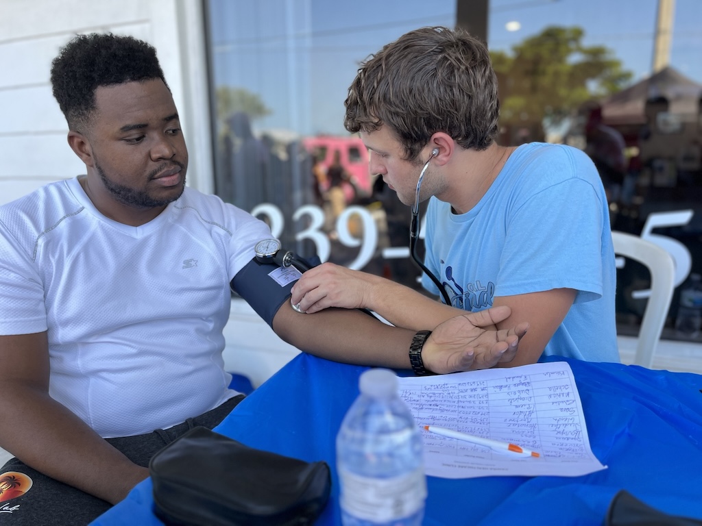 A person in a white shirt is seated while having their blood pressure checked by an FGCU student in a blue t-shirt. The student is using a stethoscope and an inflatable cuff. A blue-covered table in front of them holds a water bottle, handwritten papers and a pen. Reflections of outdoor scenery are visible in the glass windows of the building in the background. 