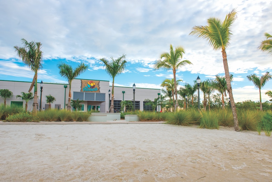  Outside view of The Boardwalk Restaurant on a sunny day with a few clouds in sight. There are about 17 palm trees, four light posts, shrubbery and beach sand in front of the restaurant. Two people can be seen in the distant background. One is wearing a red shirt.  