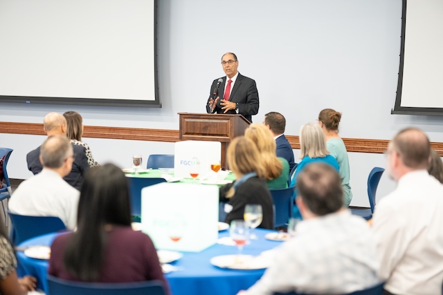 A speaker wearing a dark suit, white dress shirt, and red patterned tie stands at a podium addressing a seated audience during an indoor event. Attendees sit at round tables with blue and green tablecloths, listening as the speaker presents. Large projection screens are visible in the background.