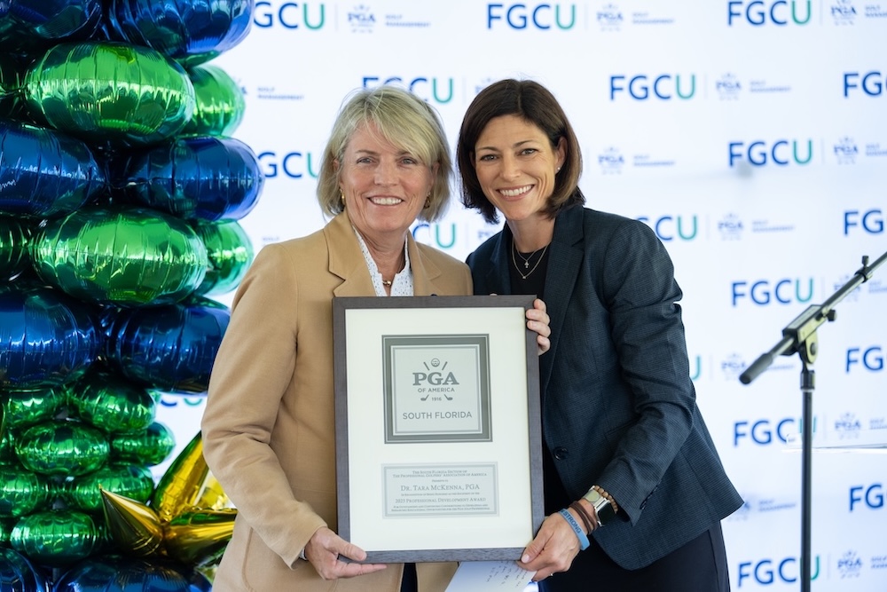 Dr. Tara McKenna, wearing a beige suit, and Meredith Schuler, wearing a gray suit, are standing together while holding a framed award. A green and blue balloon display and a white backdrop with the FGCU logo are visible behind them. 