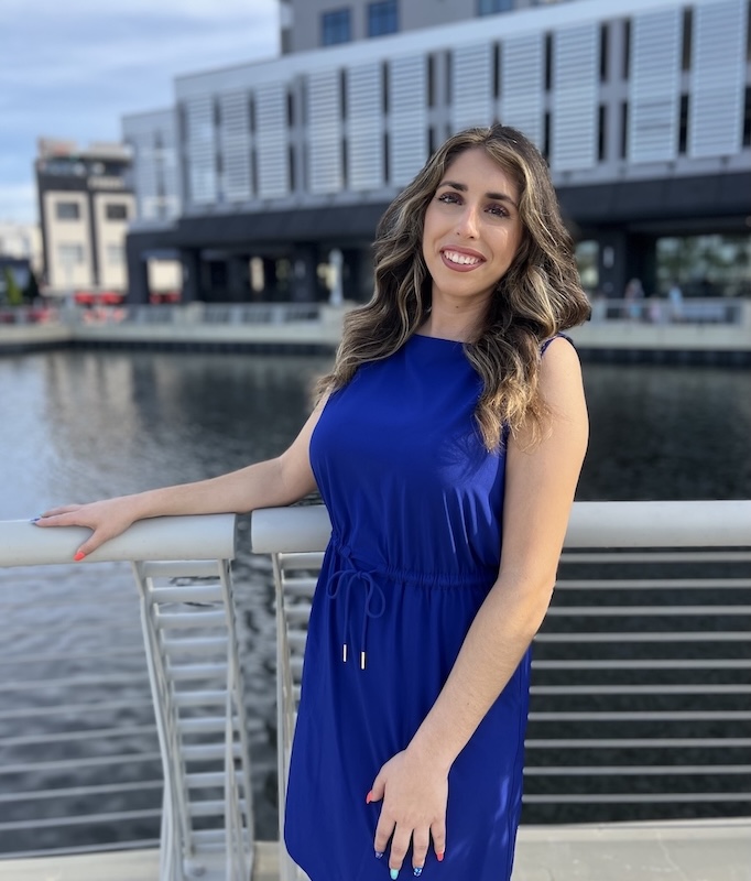 Caroline Vives-Vietri is wearing a blue dress and is posing outside in front of a body of water and buildings in the background. 