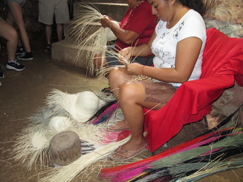 Local artisans weave traditional tan jipijapa sombreros inside a cave in Bécal, Mexico. The two women are sitting on chairs that are covered in a red blanket as they weave. The cave has a dirt floor and stairs. Students can be seen standing in the background.