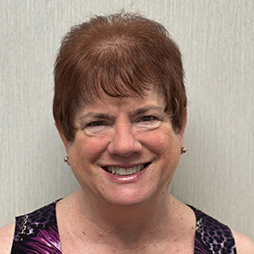 A person with short, reddish-brown hair is shown from the shoulders up, wearing a sleeveless patterned top and small stud earrings, standing against a light-colored wall. 