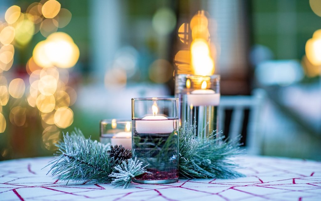 Three glass candle holders with lit white candles are arranged on a table decorated with a red geometric-patterned cloth and a green pine garland with pinecones. Warm, blurred lights glow in the background, creating a festive atmosphere.  