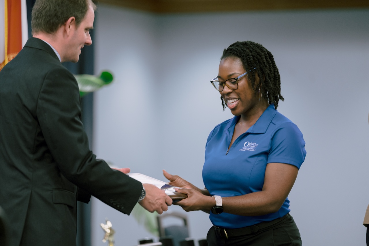 An FGCU employee, wearing glasses, a watch and a blue polo shirt with a white FGCU logo, is receiving an award from an individual wearing a black suit. There are trophies visible on a table in the background. 