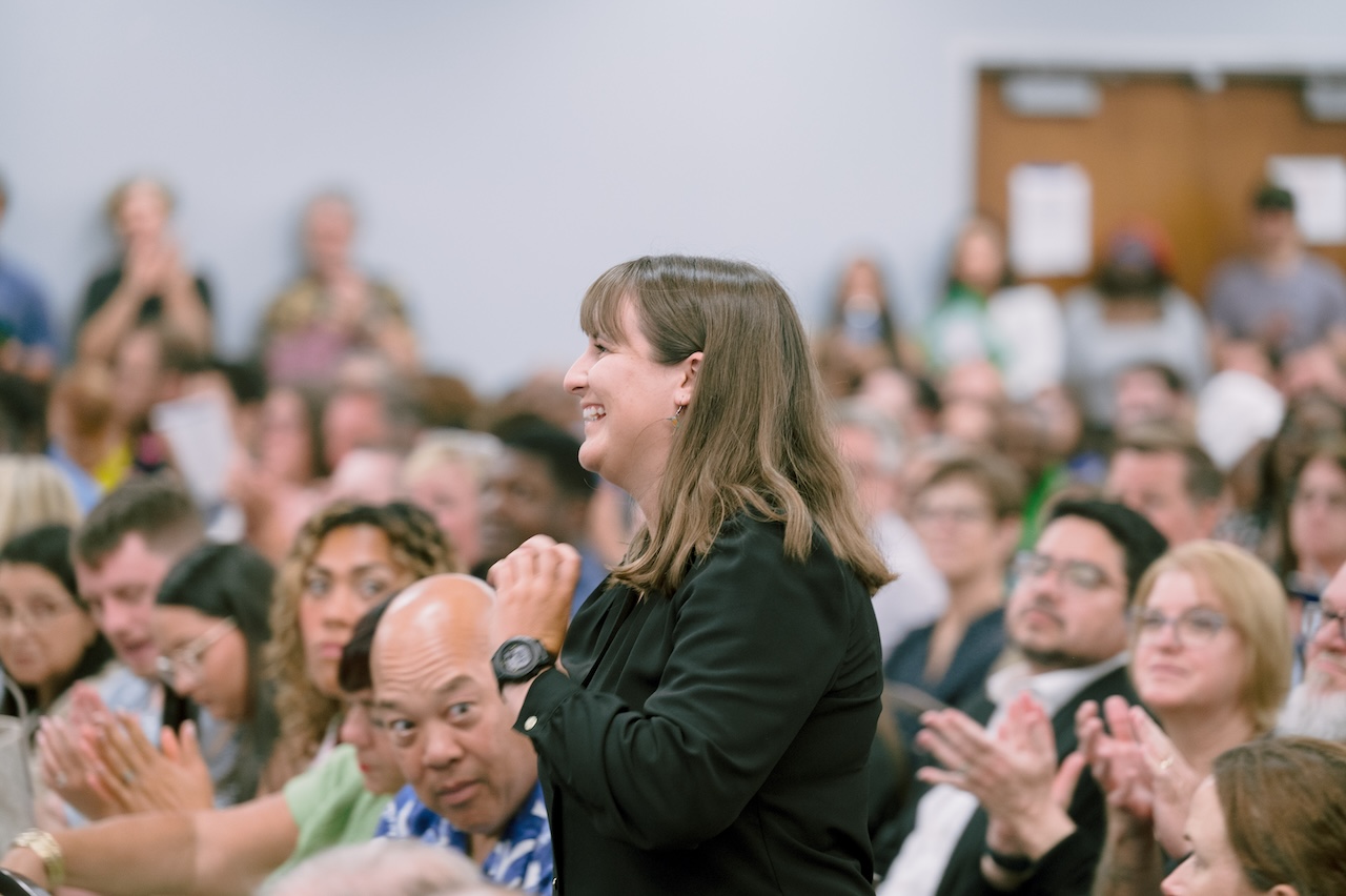 An FGCU employee is standing up among the seated crowd, and many people are clapping. She has short dark hair and is wearing a black blazer.  