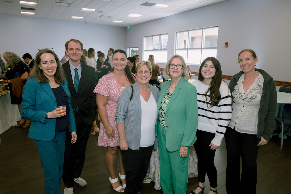 A group of FGCU employees gather for a photo at the Celebration of Excellence reception. Other employees and big windows are in the background.  The person on the left has short dark hair and is wearing a black blouse underneath a teal suit. She is holding a beverage. The next person is wearing a light blue button-down dress shirt, a green and blue tie and a black suit. The next person is wearing a pink and white floral dress and has long dark hair. The next person has short blond hair and is wearing black pants and a white blouse underneath a gray cardigan. The next person has short blonde hair, wears glasses and a green and black blouse underneath a light green suite. The next person has long dark hair and is wearing black pants and a white sweater with black stripes. The last person on the right has her hair pulled back and is wearing black pants, a cream-colored blouse with black trimming and a gray sweater on top.  