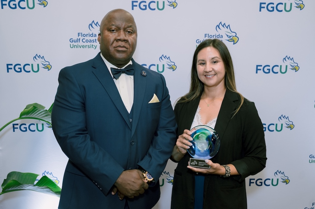 Two FGCU employees are standing in front of a backdrop with the Florida Gulf Coast University logos. The person on the left is wearing a dark blue suit, white dress shirt and a dark blue bow tie. The person on the right has dark hair and is wearing blue pants and a white blouse underneath a black blazer. She is holding an award. 
