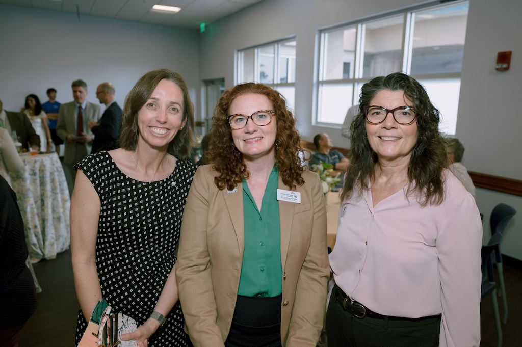 A group of three FGCU employees gather for a group photo at the Celebration of Excellence reception. The person on the left has short hair and is wearing a black dress with a white pattern. The next person has red curly hair and is wearing black pants and a green blouse underneath a beige blazer. The last person on the right has short dark hair, is wearing glasses, black pants and a light pink blouse. 