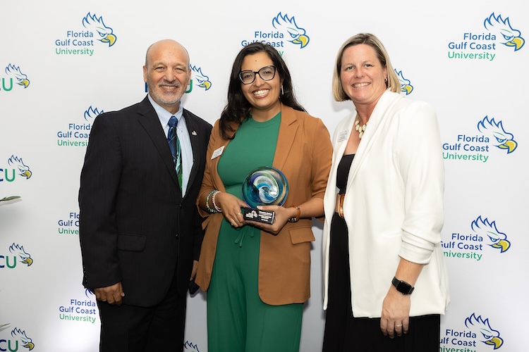Vice Provost Paul Dosal, an FGCU employee, and Tara McKenna, Director of the Golf Management, stand in front of a FGCU step-and-repeat backdrop. The person in the middle holds a glass award.