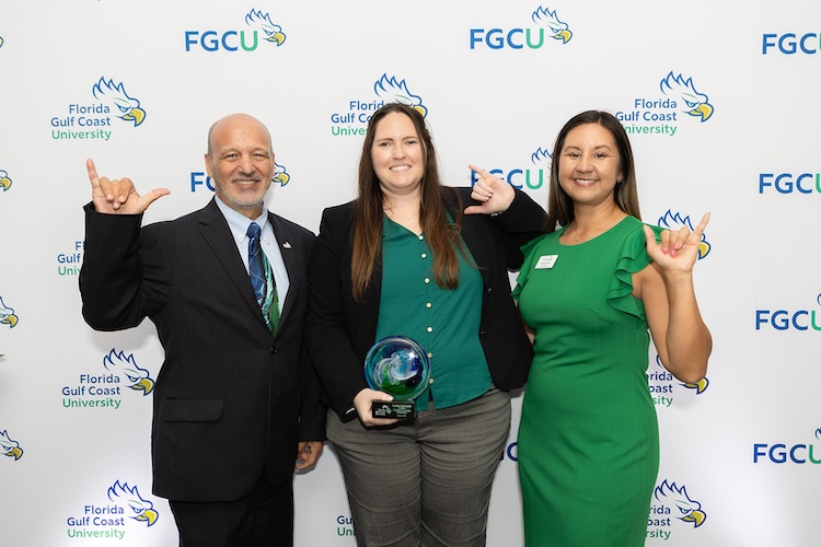 Vice Provost Paul Dosal and two FGCU employees stand in front of a FGCU step-and-repeat. The person in the middle holds a blue and green glass award while all three make the Wings Up sign.