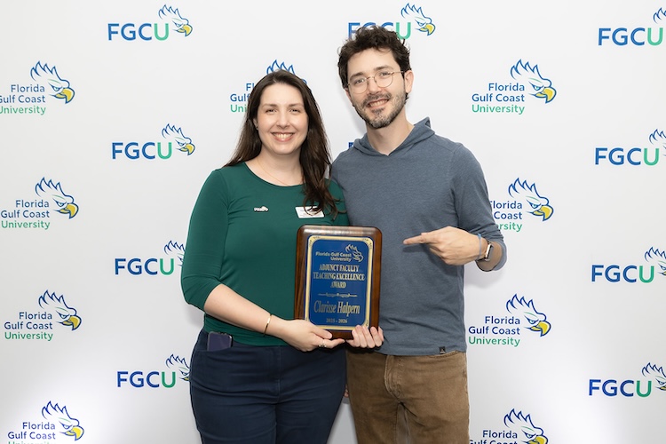 Two FGCU employees pose for a photo while the woman to the left is holding up a blue plaque award.