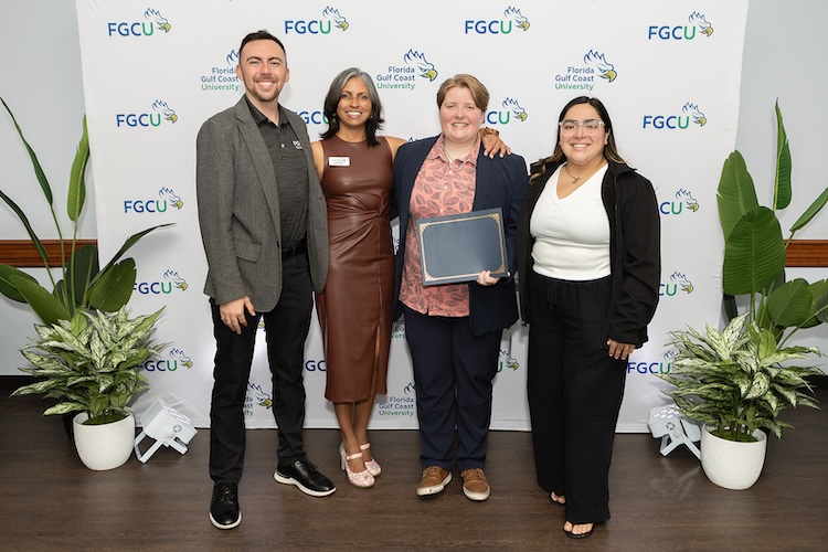 A group of four FGCU employees pose for a photo in front of a white FGCU step-and–repeat. The third person to the right is holding up a blue and gold certificate folder.