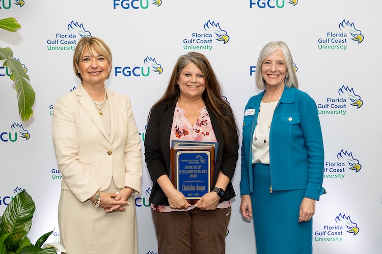 FGCU President Aysegul Timur (dressed in a cream-colored suite) stands in front of an FGCU step-and-repeat with the award recipient, (dressed in a pink blouse, black cardigan and brown pants) and Provost Debbie Thorne (dressed in a blue skirt and jacket)