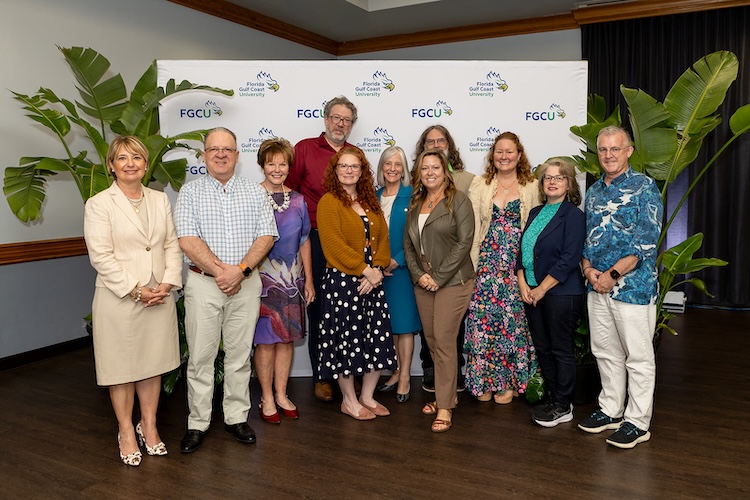 A group of FGCU employees, including FGCU President Aysegul Timur, pose together in front of a white FGCU step-and-repeat inside the Cohen Student Union Ballroom.