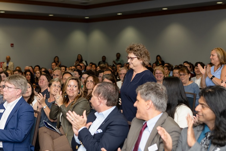 A FGCU employee stands in the middle of a seated audience in the Cohen Student Union Ballroom while many attendees around her applaud.