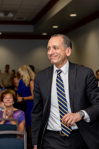 An FGCU employee (dressed in a black suit, white shirt and black and white striped tie) walks past the crowd.