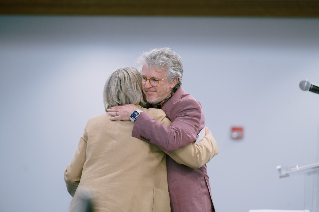 Two individuals are hugging.  One person is wearing a beige jacket and has short, light-colored hair. The other person is wearing a pinkish jacket and also has grey hair. There is a microphone on a stand to the right side.