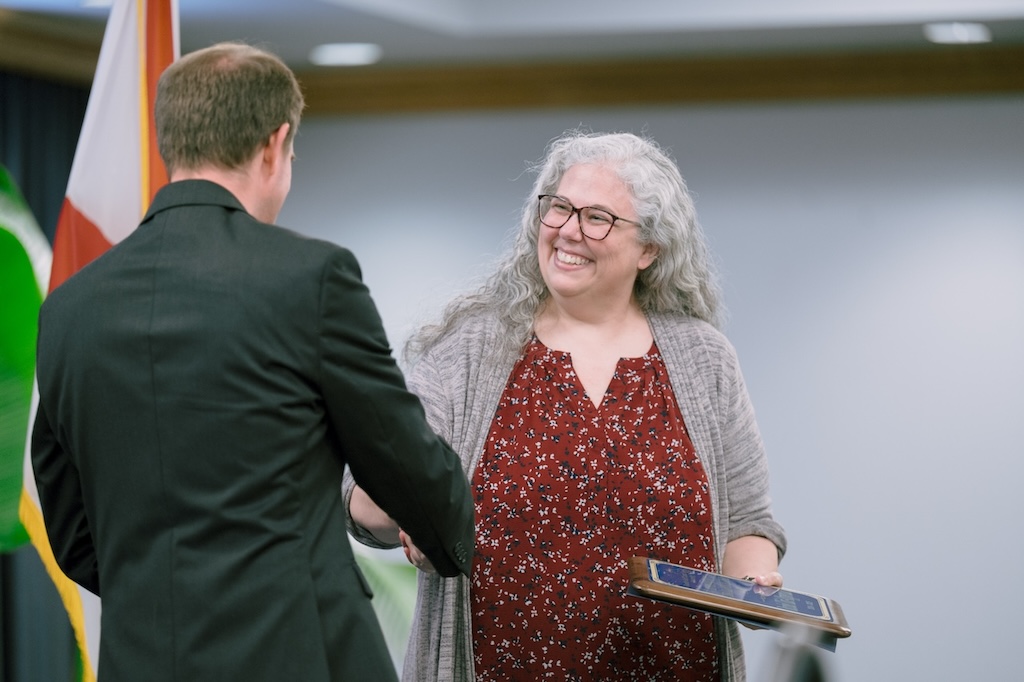 An FGCU employee, who has long grey curly hair and is wearing a red floral blouse underneath a gray cardigan, is receiving an award and shaking the hand of an individual wearing a black blazer. 