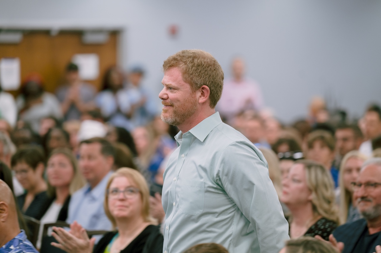 An audience member is standing up among the seated crowd. He has blond hair and is wearing a light blue button-down shirt.  