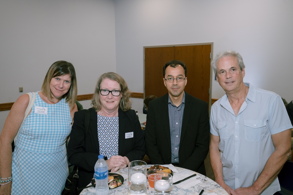 A group of four FGCU employees pose for a photo at the Celebration of Excellence reception. The person on the far left has short blond hair and is wearing a light blue checkered dress. The person next to her has short blond wavy hair and is wearing glasses, a blue-patterned blouse and a black blazer. The next person has black hair and is wearing glasses, a blue button-down shirt and a black blazer. The fourth person has short grey hair, and he is wearing a light blue bottom-down shirt.  