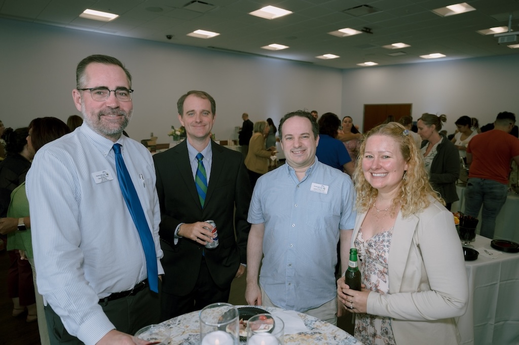 A group of four FGCU employees are posing for a group photo at the Celebration of Excellence reception. Tables and chairs are in the background. The individual on the far left is wearing glasses, a light blue button-down dress shirt and a blue tie. The next person is wearing a blue button-down dress shirt, a green and blue tie and a black blazer. He is holding a diet Pepsi can. The third person is wearing a short sleeve button-down shirt with his FGCU name tag. The last person is wearing a floral dress underneath a beige blazer. She has curly blond hair.  