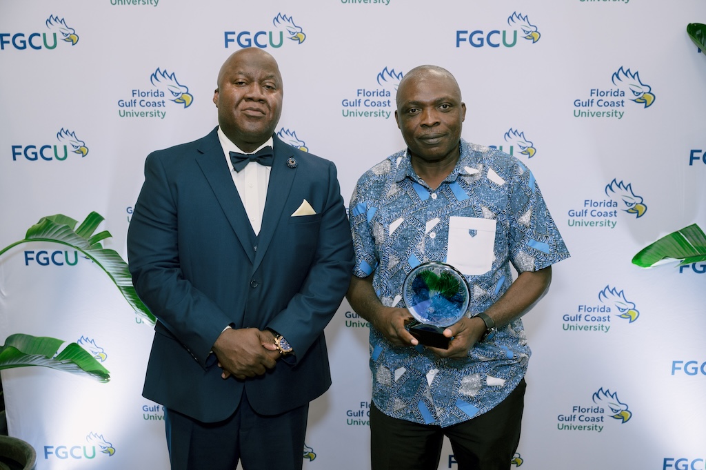 Two FGCU employees are standing in front of a backdrop with the Florida Gulf Coast University logos. The person on the left is wearing a dark blue suit, white dress shirt and a dark blue bow tie. The person on the right is wearing a blue short-sleeved shirt with white patterns and is holding an award.  