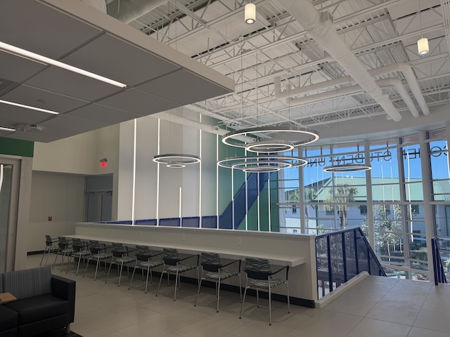A bright, modern interior space with high ceilings, circular pendant lights, and tall windows. A long counter with chairs lined on the left side, and a staircase with a blue railing descends near the large glass wall overlooking palm trees outside.