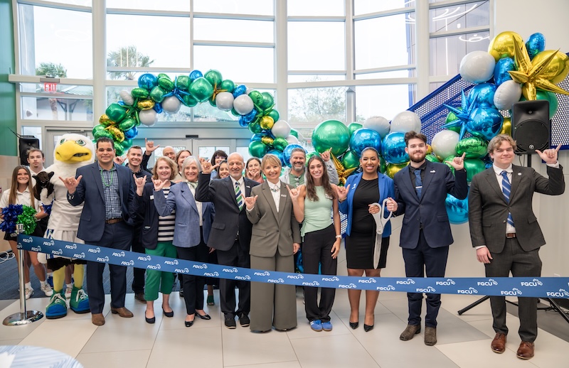 A large group of FGCU employees and students stand together inside the Cohen Student Union main entrance at a ribbon‑cutting event while doing the Wings Up sign. A blue, green, white, and gold balloon display is visible behind them.