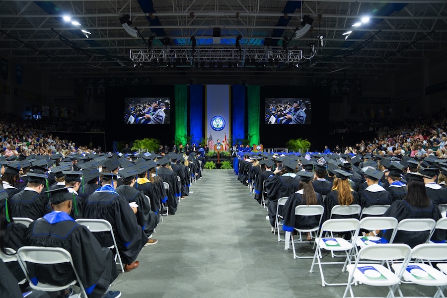 View of an FGCU commencement ceremony at Alico Arena. Graduates are seated on the main floor while FGCU President Aysegul Timur gives a speech. Guests can be seen seated on the side bleachers. 