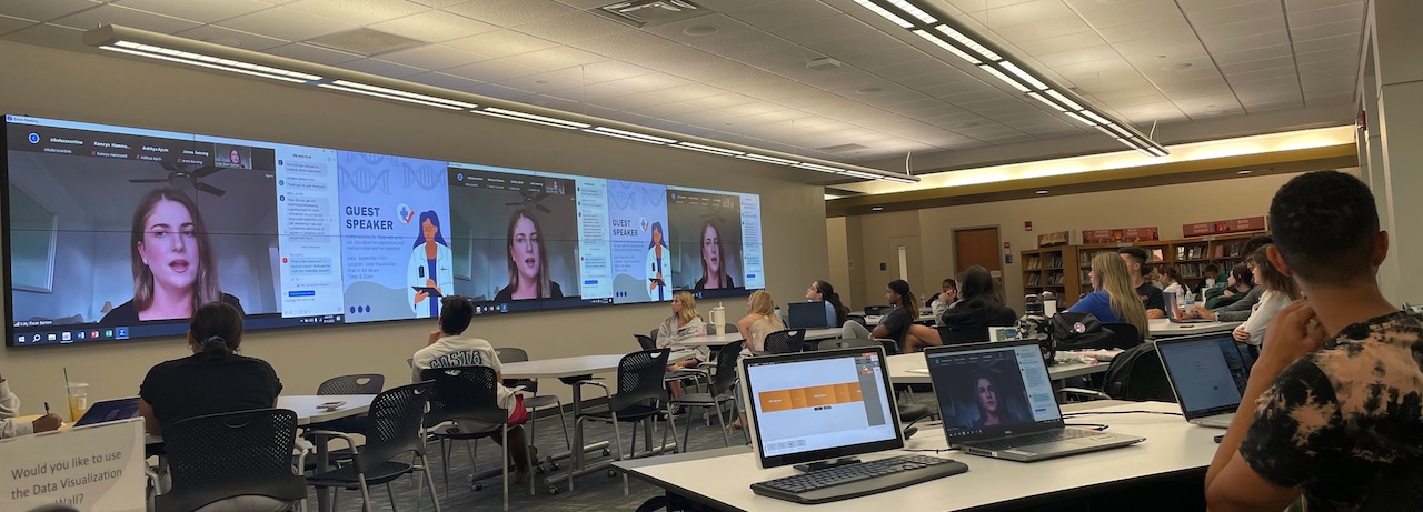 People are seated at tables with laptops, watching a virtual guest speaker displayed on multiple large screens (visualization wall) at the front of the room. The screens show the speaker’s face and the text 'GUEST SPEAKER' alongside an illustration of a person in a lab coat. The room features contemporary lighting and a hybrid learning setup that enables remote interaction with in-person attendees.  