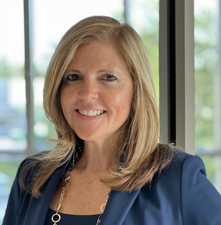 Headshot of Dr. Eileen DeLuca. She has short blond hair and is wearing a blue blazer and necklace. Windows can be seen in the background.