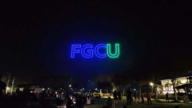 Dozens of brightly lit drones form the letters “FGCU” in blue and green against the night sky, while a crowd of people gathers below to watch the display on a grassy campus area known as the library lawn. 