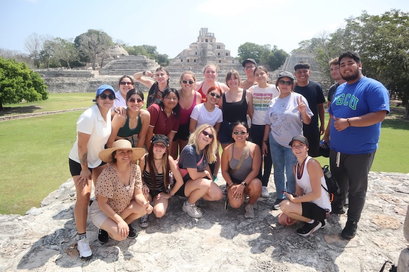 FGCU students, a staff member, and students from Instituto Campechano are posing for a photo together outdoors in front of Edzná, a Mayan archaeological site in Mexico. The group wears sunglasses and athletic clothes as they stand on a rocky surface with grass surrounding them. 