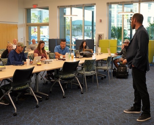 Veterans Florida Entrepreneurship Program participants are sitting down on green chairs in front of long rectangular tables taking notes during the “Selling and Leadership Effectiveness” workshop lead by Jason Cutter, author of “Selling with Authentic Persuasion.” The people sitting have drinks, computers, and bags on top of the tables. There are also outlets on the tables. Floor-to-ceiling windows can be seen on the right side of the room as well as light green and gray furniture. The speaker is wearing a dark gray suit and is facing the participants.  