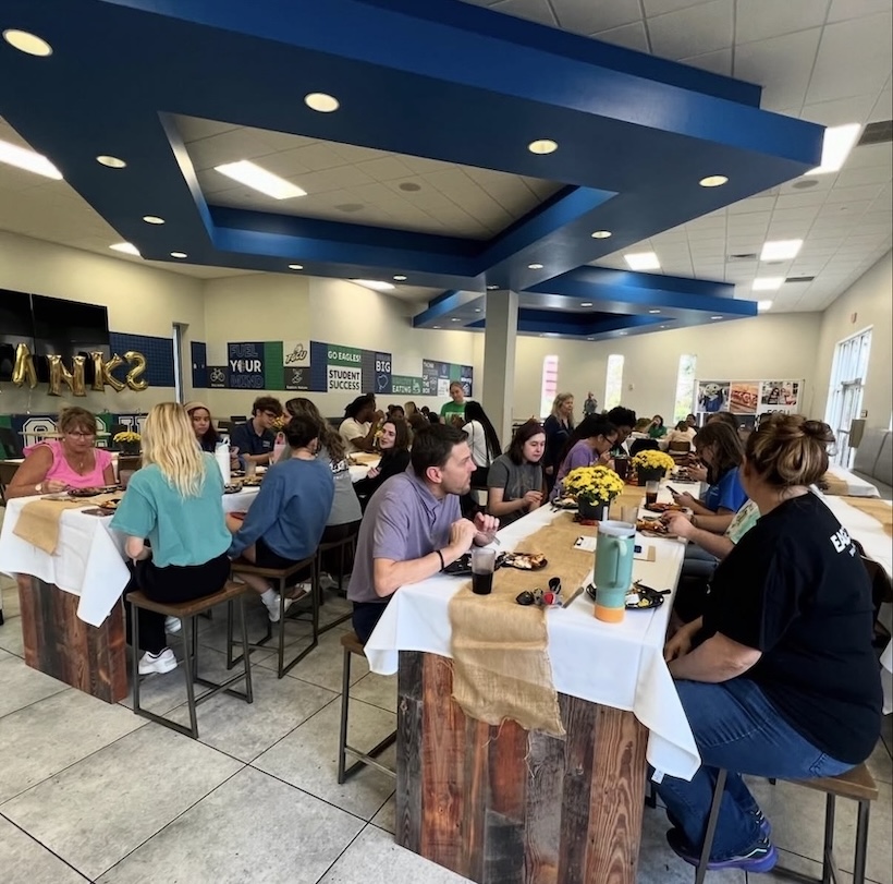 FGCU faculty and staff are at Sovi Dining, eating lunch on long wooden tables with white tablecloths and burlaps fabric on top. Yellow flowerpots, food, and drinks are on the tables.  