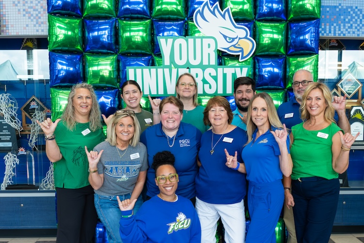 A group of FGCU employees dressed in blue and green clothing pose in front of a backdrop made up of green and blue balloons, the FGCU bird logo and the wording “Your University” in white and outlined in green. They are giving the “wings up” salute.