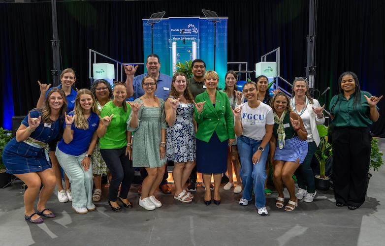 A group of 15 employes pose together with President Aysegul Timur in front of a stage inside Alico Arena. All are dressed in green and blue clothing and are doing the “wings up” salute. The stage backdrop includes black curtains, blue lighting, two LED cubes displaying the FGCU logo, and a central podium with a blue to green ombre backdrop. Potted plants surround the stage area.