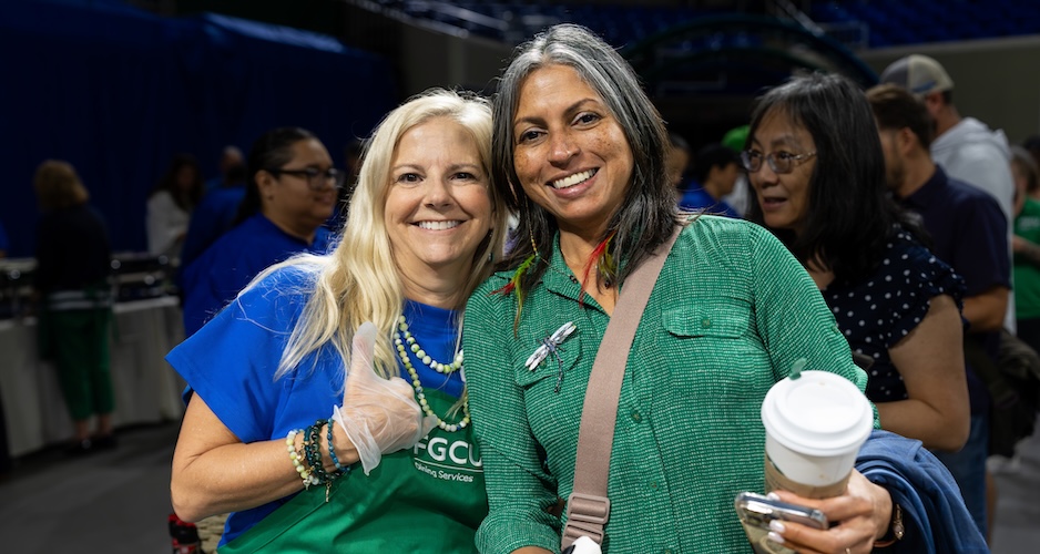 Two FGCU employees pose together for a photo. The employee on the lef gives a thumbs-up while wearing a green FGCU Dining Services apron and blue t-shirt underneath. The employee on the right wears green long-sleeve blouse and is holding a coffee cup in her hand.