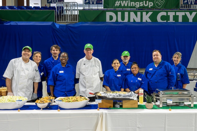 Dining services staff pose behind a serving table filled with prepared dishes. Team members wear blue and white uniforms and chef coats, with serving equipment and labeled food items arranged across the table. Some are also wearing green caps.