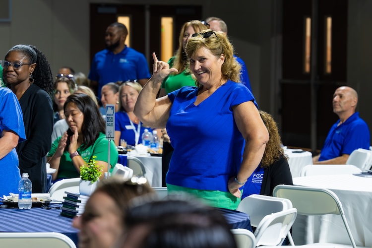 An FGCU employee is standing up beside her table and makes the FGCU “wing up” salute while facing the camera. She is wearing a blue blouse and green pants. Other event participants are seated and standing throughout the background inside Alico Arena.