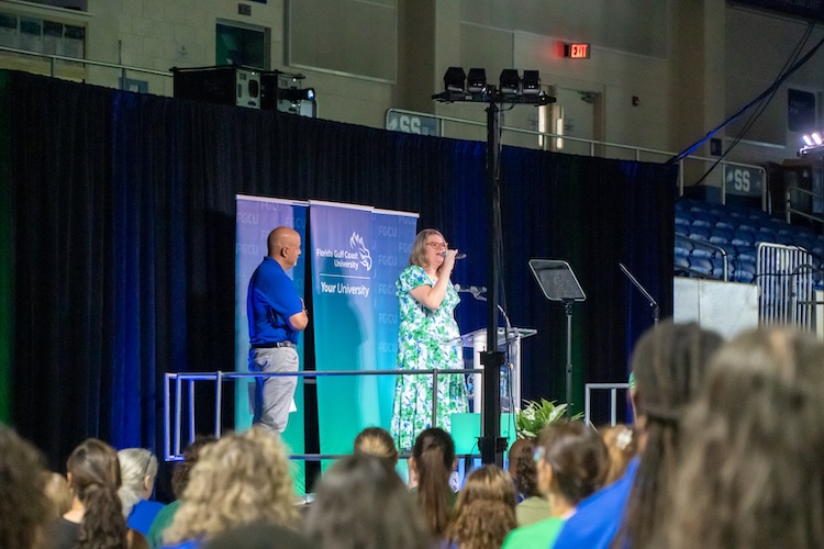 A stage is set up inside Alico Arena with audience members seated in front. On the stage, an FGCU employee is singing the National Anthem. She is wearing a green and white dress. Another employee is standing next to her watching her sing. He is wearing a blue polo and grey pants. FGCU-branded banners are displayed behind them, and a teleprompter is visible on stage.